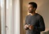 A man with short curly dark hair and a charcoal sweater standing near a window, holding a coffee cup and looking out with a pensive, reflective expression during warm late afternoon light.
