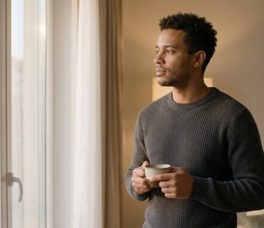 A man with short curly dark hair and a charcoal sweater standing near a window, holding a coffee cup and looking out with a pensive, reflective expression during warm late afternoon light.
