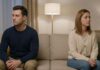A man and woman sitting at opposite ends of a long living room couch in soft evening lamp light, looking away thoughtfully to capture emotional distance.