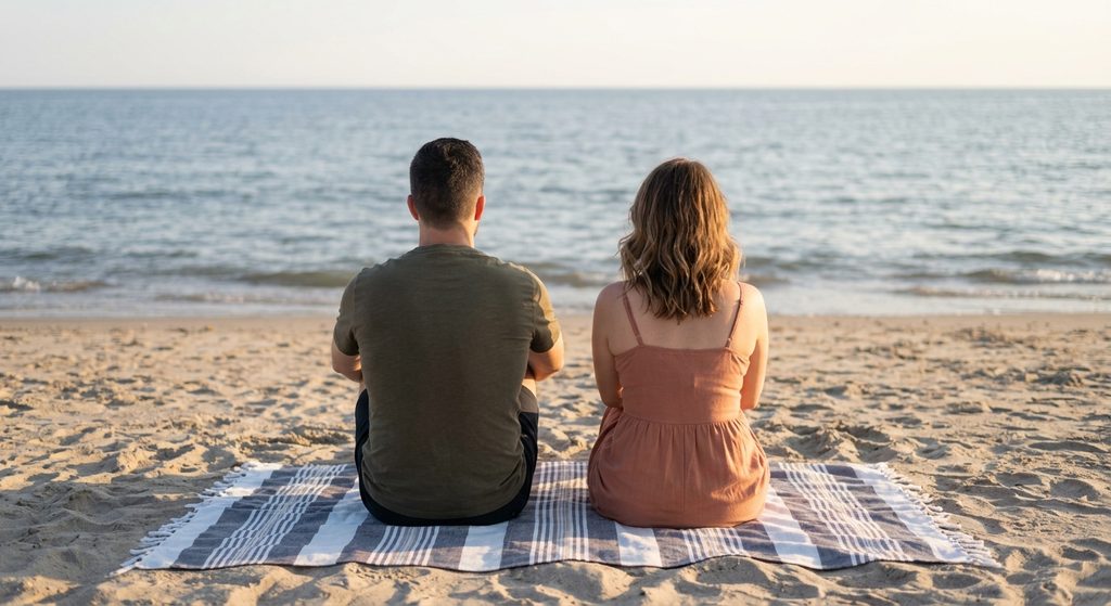 The couple, featuring the man and woman from previous images, sitting side-by-side on a beach blanket, staring straight ahead at the ocean in total, heavy silence with a noticeable physical and emotional gap between them.
