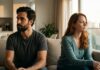 The dark-haired man and auburn-haired woman from previous images sitting on opposite ends of a contemporary neutral-toned sofa in warm evening natural light, physically close but emotionally distant, lost in reflection.