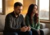 A couple sits on a modern living room couch, physically close but emotionally disconnected—the woman with auburn hair looks thoughtfully out the window while the man looks down with a worried expression, in warm evening light.