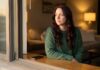 A thoughtful woman with long auburn hair and a forest-green sweater sits on a window sill looking outside during golden hour, lost in reflection, while her phone rests face down beside her.