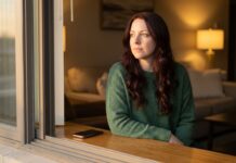 A thoughtful woman with long auburn hair and a forest-green sweater sits on a window sill looking outside during golden hour, lost in reflection, while her phone rests face down beside her.