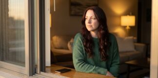 A thoughtful woman with long auburn hair and a forest-green sweater sits on a window sill looking outside during golden hour, lost in reflection, while her phone rests face down beside her.