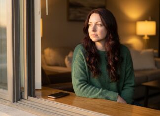 A thoughtful woman with long auburn hair and a forest-green sweater sits on a window sill looking outside during golden hour, lost in reflection, while her phone rests face down beside her.